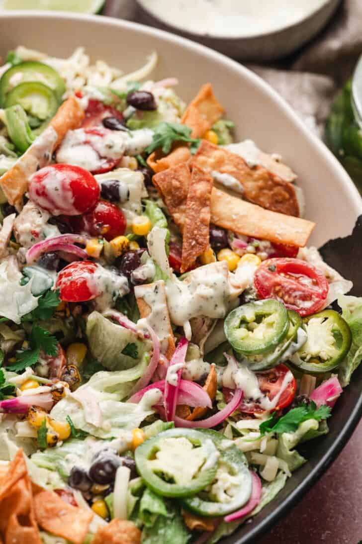 Close-up of a southwest salad with chicken, showing shredded chicken, black beans, corn, cherry tomatoes, jalapeño slices, pickled red onion, and crispy tortilla strips, topped with a drizzle of creamy ranch dressing.