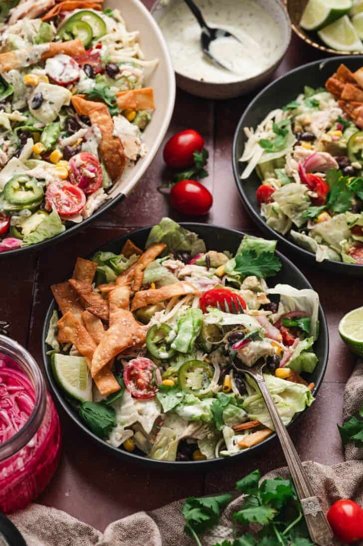 Overhead view of three bowls of southwestern chicken salad arranged on a table with cherry tomatoes, lime wedges, a jar of pickled red onions, and a bowl of ranch dressing, with one bowl in the foreground being eaten with a fork.