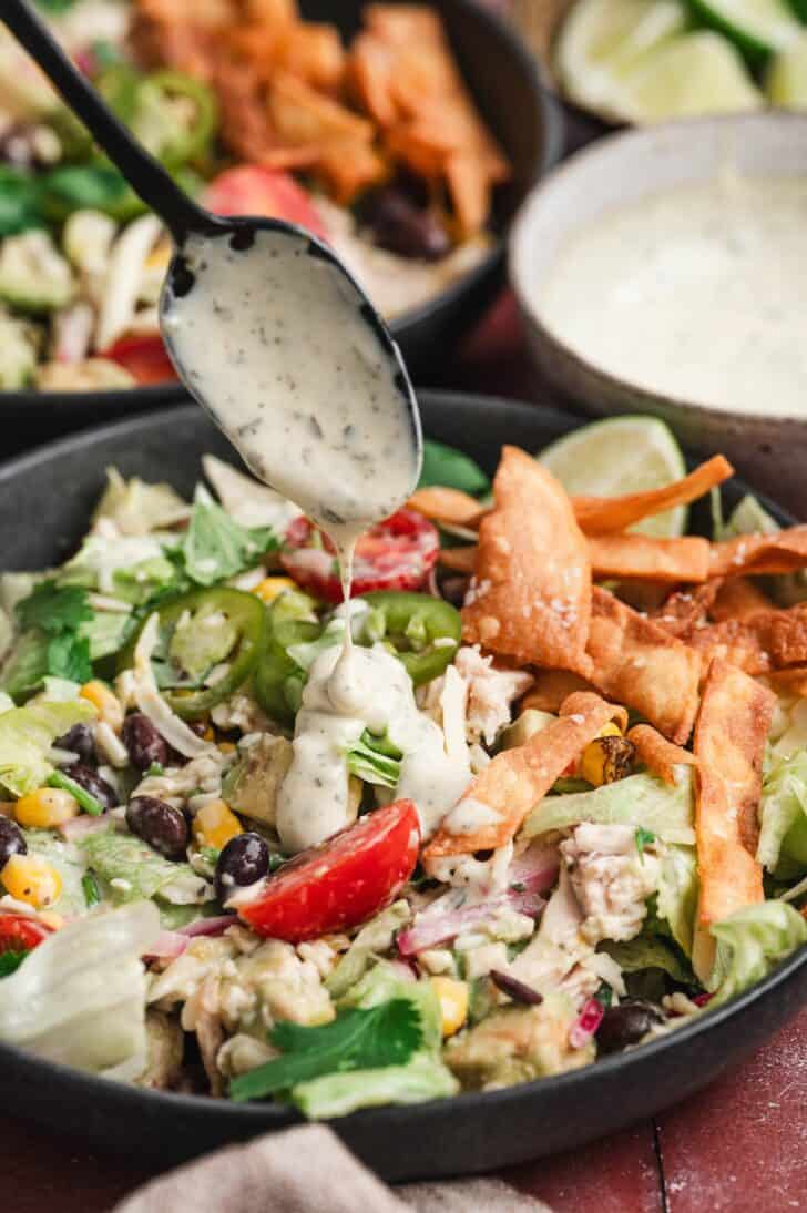 Close-up of ranch dressing being spooned over a southwestern salad with chicken, topped with black beans, corn, cherry tomatoes, jalapeño slices, and crispy tortilla strips.