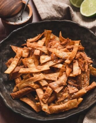Overhead view of a bowl piled with fried tortilla strips, sprinkled with sea salt and served with lime halves and a small dish of salt.