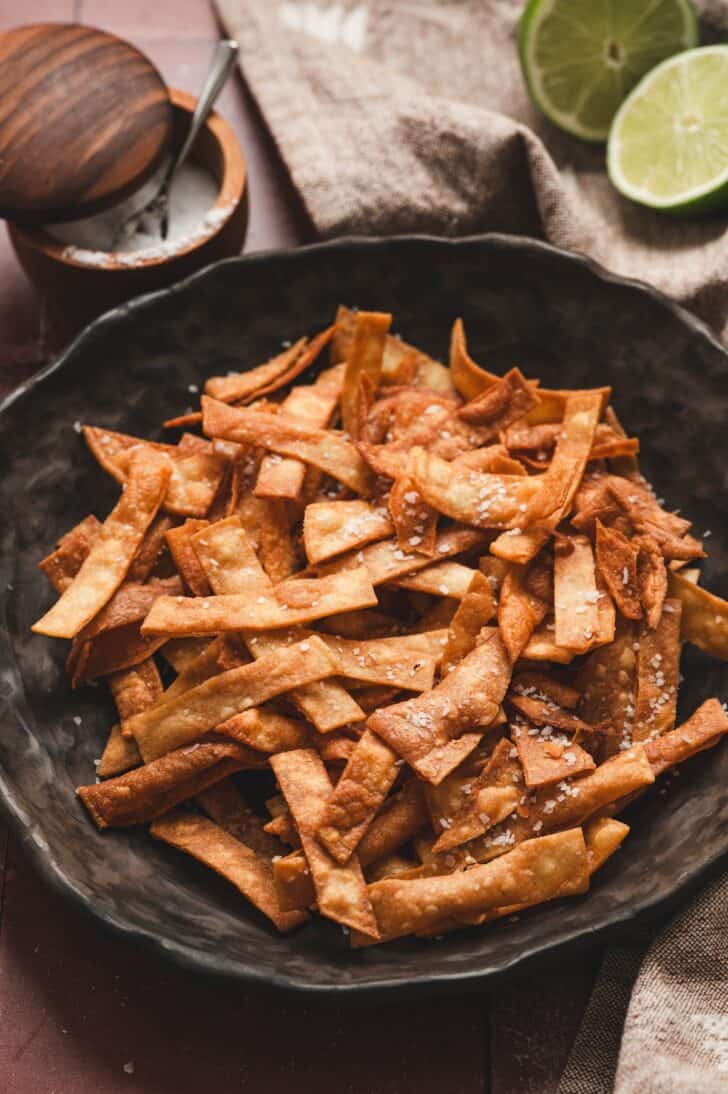 Overhead view of a bowl piled with fried tortilla strips, sprinkled with sea salt and served with lime halves and a small dish of salt.