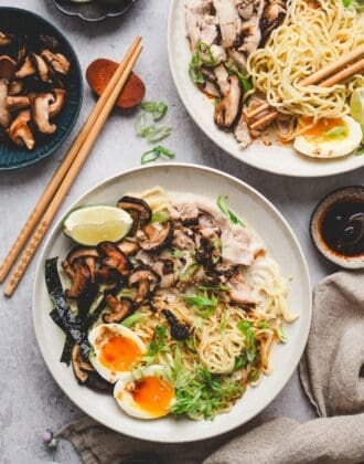 Two plates of chicken ramen recipe topped with sliced chicken, sautéed shiitake mushrooms, soft-boiled eggs, scallions, and lime wedges