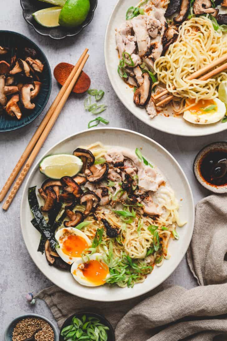 Two plates of chicken ramen recipe topped with sliced chicken, sautéed shiitake mushrooms, soft-boiled eggs, scallions, and lime wedges