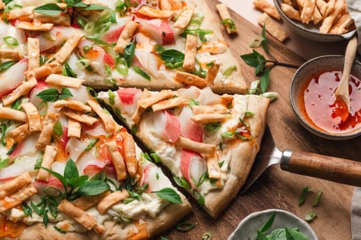 Close-up of sliced crab rangoon flatbread topped with imitation crab, melted mozzarella, green onions, Thai basil, and crispy wonton strips, drizzled with sweet chili sauce.
