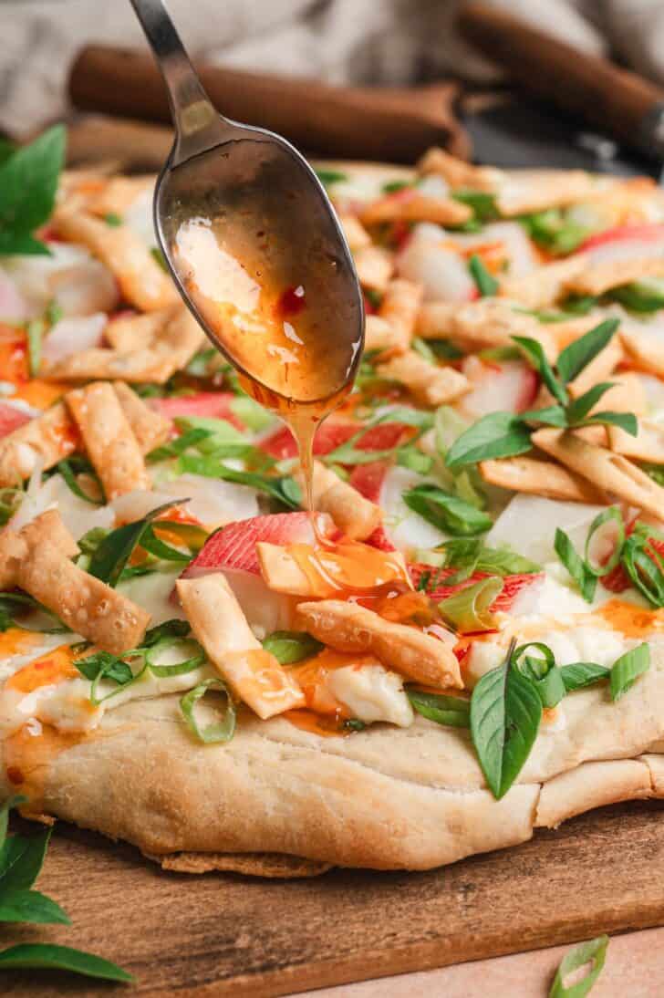 Close-up of a crab pizza recipe with a spoon drizzling sweet chili sauce over wonton strips, Thai basil, green onions, and imitation crab.