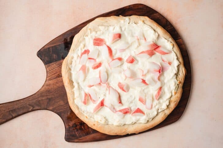 Overhead view of pizza dough topped with a cream cheese mixture and imitation crab pieces on a wooden pizza peel, ready to bake.