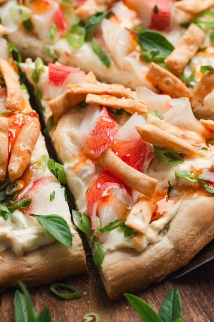 Close-up of sliced crab rangoon flatbread topped with imitation crab, melted mozzarella, green onions, Thai basil, and crispy wonton strips, drizzled with sweet chili sauce.