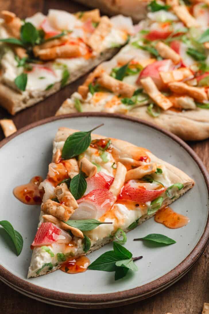 A slice of crab rangoon pizza on a plate, topped with imitation crab, melted mozzarella, green onions, Thai basil, and crispy wonton strips, drizzled with sweet chili sauce, with more slices in the background.