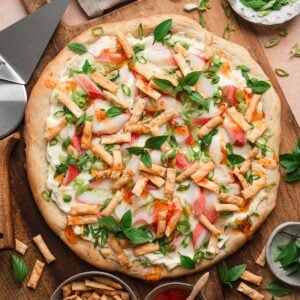 Overhead view of a whole crab rangoon pizza on a wooden board, topped with imitation crab, melted mozzarella, green onions, Thai basil, and crispy wonton strips, drizzled with sweet chili sauce.