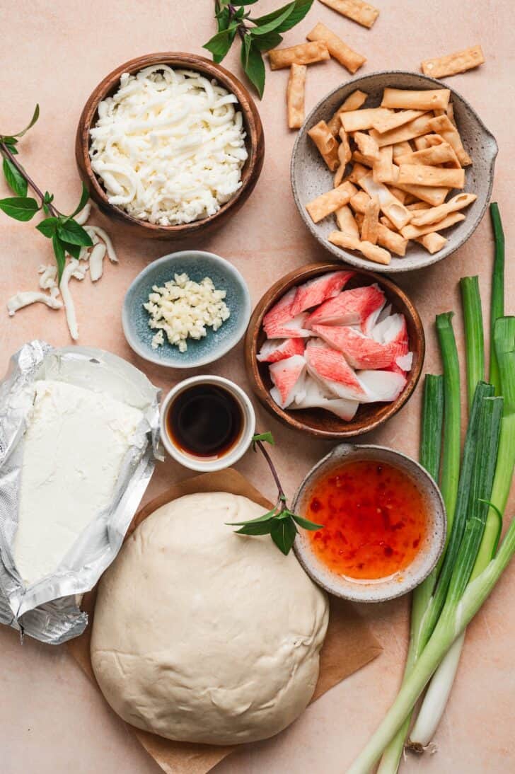 Flat-lay of ingredients for crab rangoon pizza, including shredded mozzarella cheese, wonton strips, minced garlic, imitation crab meat, cream cheese, Worcestershire sauce, pizza dough, sweet chili sauce, and green onions.