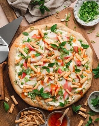 Overhead view of a whole crab rangoon pizza recipe on a wooden board, topped with imitation crab, melted mozzarella, green onions, Thai basil, and crispy wonton strips, drizzled with sweet chili sauce, with bowls of green onions, wonton strips, and chili sauce alongside.