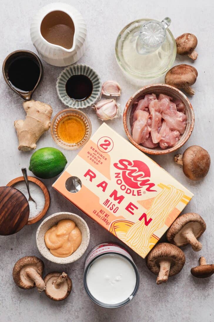 Ingredients for creamy chicken ramen laid out, including raw chicken, shiitake mushrooms, miso paste, coconut milk, soy sauce, ginger, garlic, and a box of ramen noodles