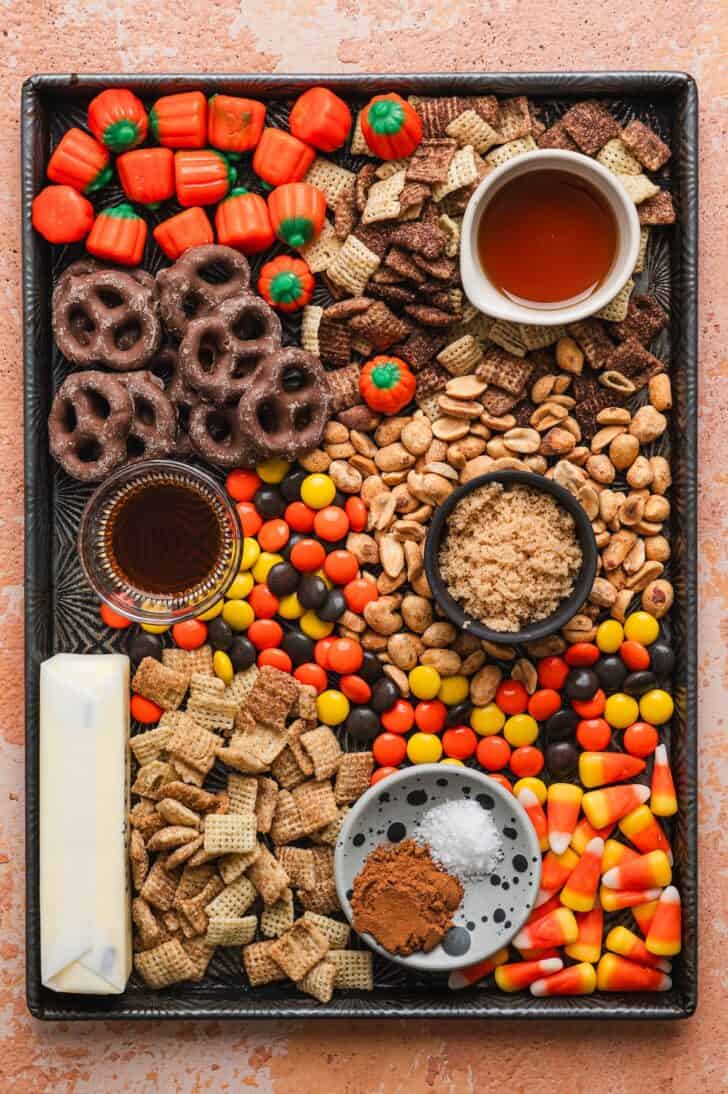 Ingredients for halloween chex mix recipe arranged on a sheet pan, including Chex cereal, chocolate-covered pretzels, candy corn, mellowcreme pumpkins, Reese's Pieces, peanuts, butter, and maple syrup