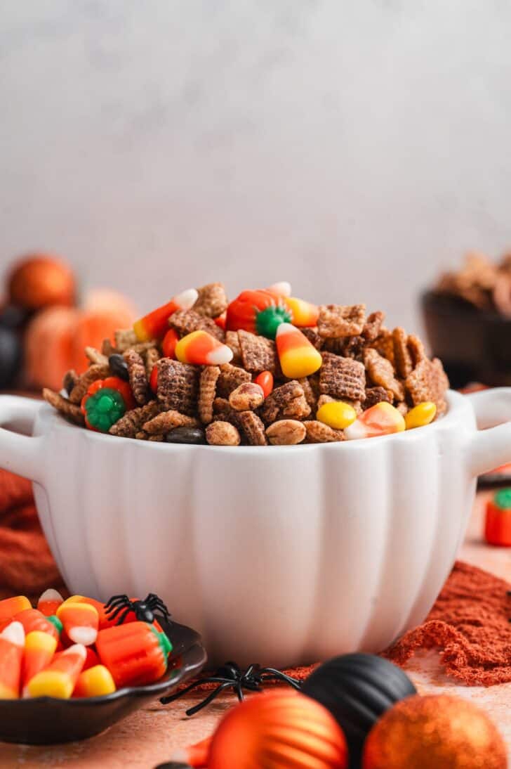 Pumpkin-shaped bowl filled with halloween chex mix recipe, with a small dish of candy corn and mellowcreme pumpkins alongside