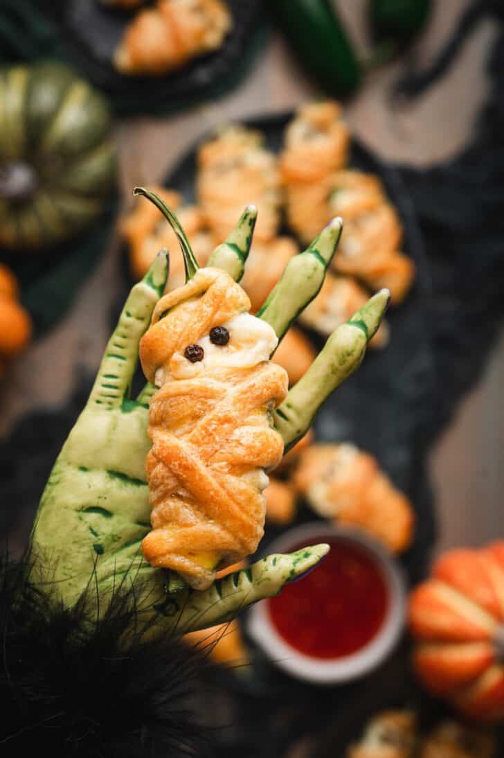 Close-up of a halloween jalapeno popper shaped like a mummy, wrapped in golden pastry strips with peppercorn eyes, resting on a spooky hand-shaped dish next to a bowl of red dipping sauce, with more poppers and mini pumpkins blurred in the background.