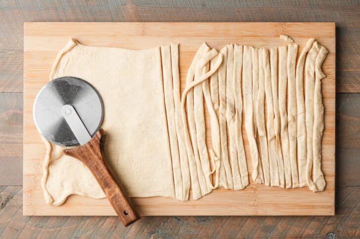 Overhead view of a sheet of crescent dough cut into thin strips on a wooden cutting board with a pizza cutter.