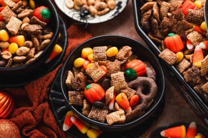 Three bowls of halloween party mix filled with cereal, candy corn, mellowcreme pumpkins, peanuts, and chocolate-covered pretzels