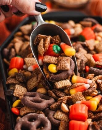 Scoop lifting a serving of halloween snack mix loaded with candy corn, a mellowcreme pumpkin, and a chocolate-covered pretzel