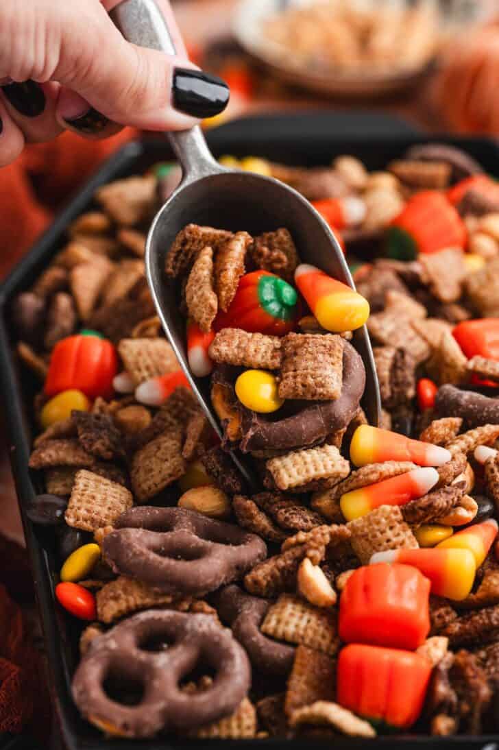 Scoop lifting a serving of halloween snack mix loaded with candy corn, a mellowcreme pumpkin, and a chocolate-covered pretzel