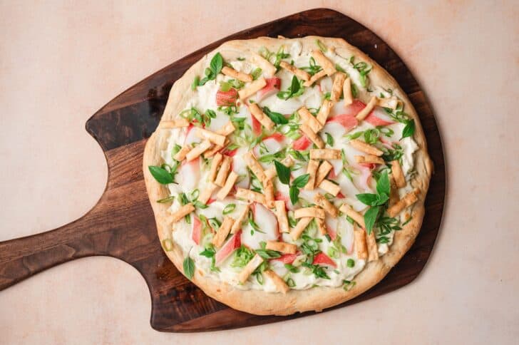 Overhead view of an imitation crab pizza on a wooden peel, topped with imitation crab, melted mozzarella, green onions, and Thai basil.
