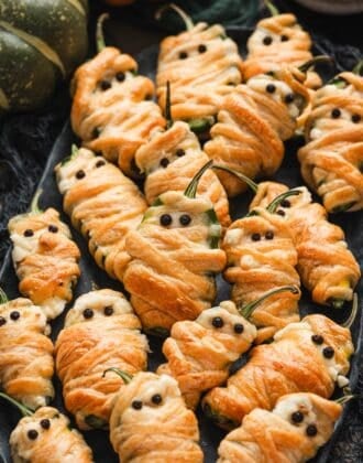 Overhead view of a platter piled with jalapeno mummy poppers, wrapped in golden pastry with peppercorn eyes, surrounded by mini pumpkins and black cheesecloth for a Halloween spread.