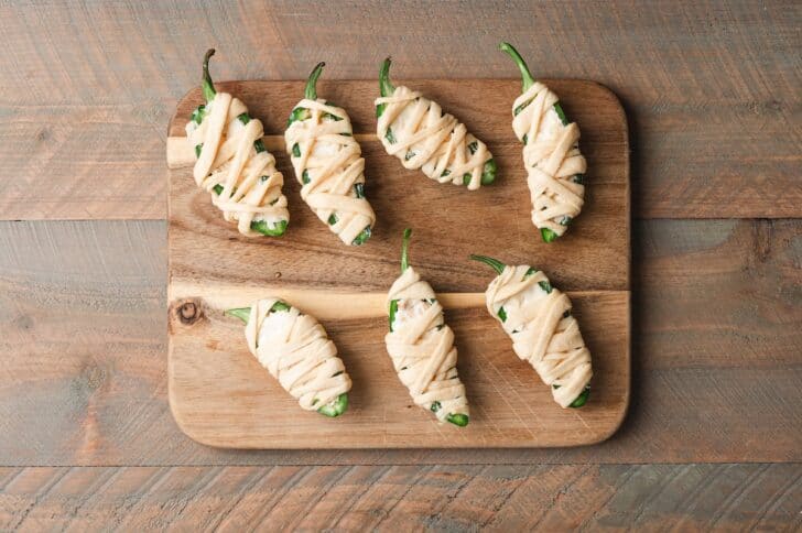 Overhead view of jalapeño pepper halves wrapped in strips of dough on a wooden cutting board, before baking.