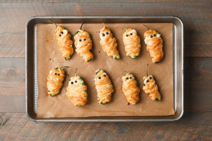 Overhead view of baked mummy jalapeño poppers with peppercorn eyes, arranged in rows on a parchment-lined baking sheet.