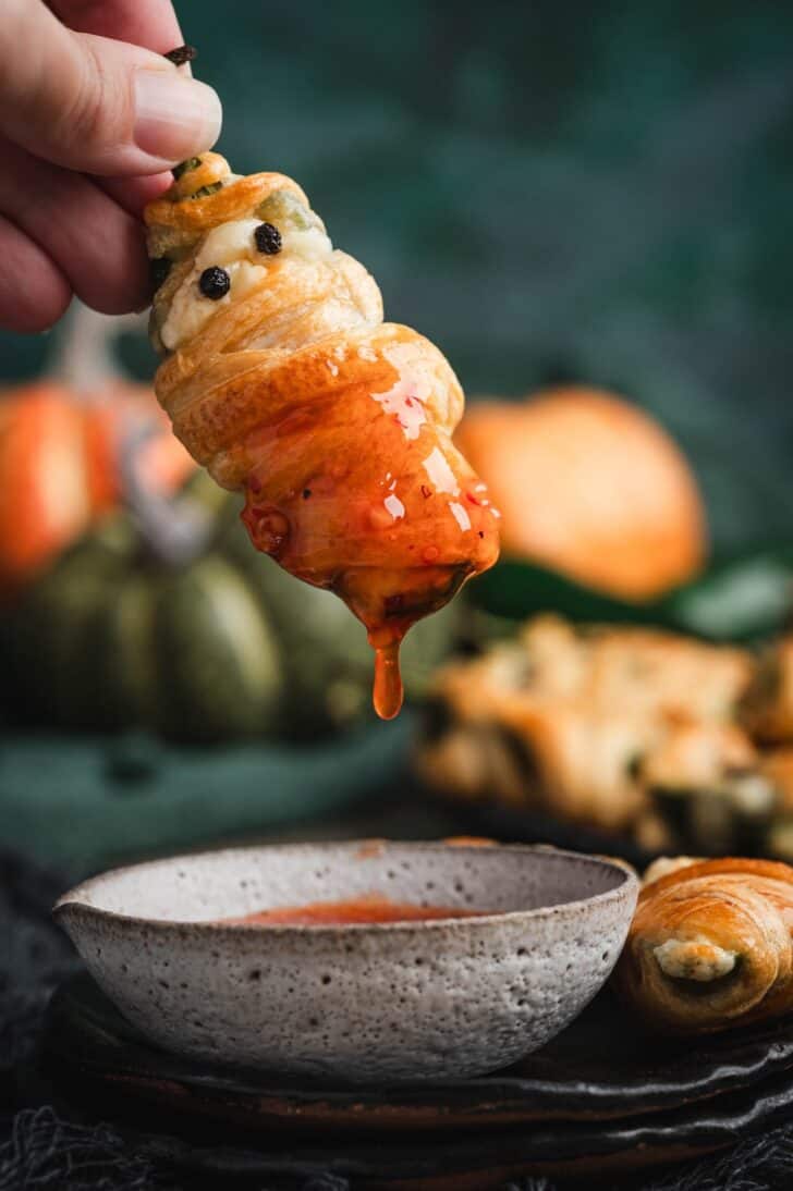 Close-up of a hand holding a jalapeno popper mummy dripping with sweet chili sauce over a small bowl, with more poppers and mini pumpkins blurred in the background.