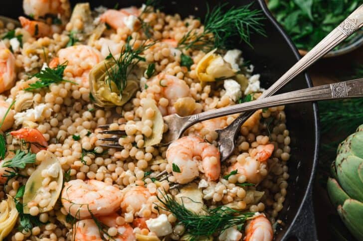 Close-up of a lemon shrimp couscous skillet with shrimp, artichokes, and pearl couscous, garnished with crumbled feta, fresh dill, and parsley, with two forks digging in.