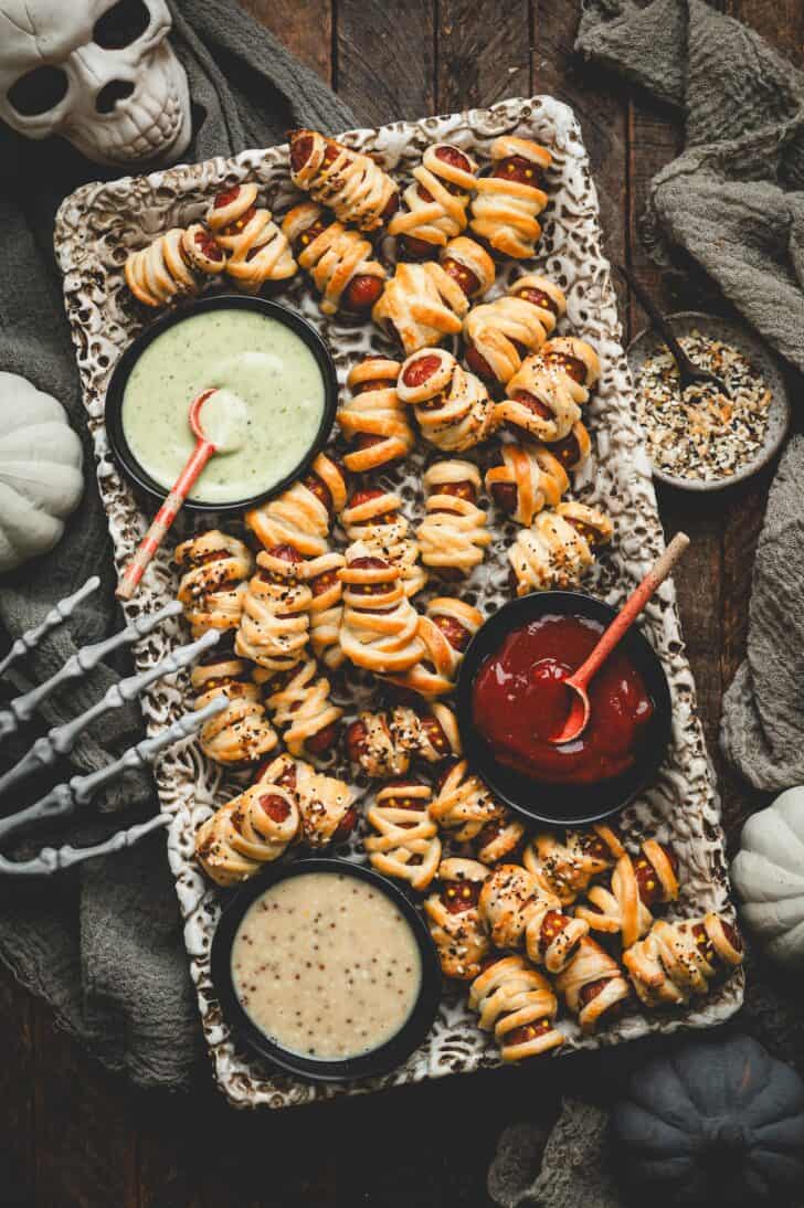 Overhead platter of lil smokies pigs in a blanket arranged with dipping sauces, a ceramic skull, and a skeleton hand