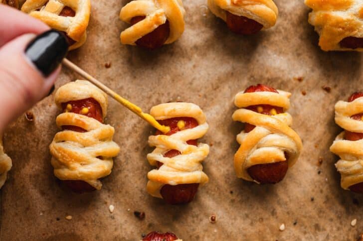 Mustard being dotted onto baked lil smokies pigs in a blanket with a toothpick to create mummy eyes