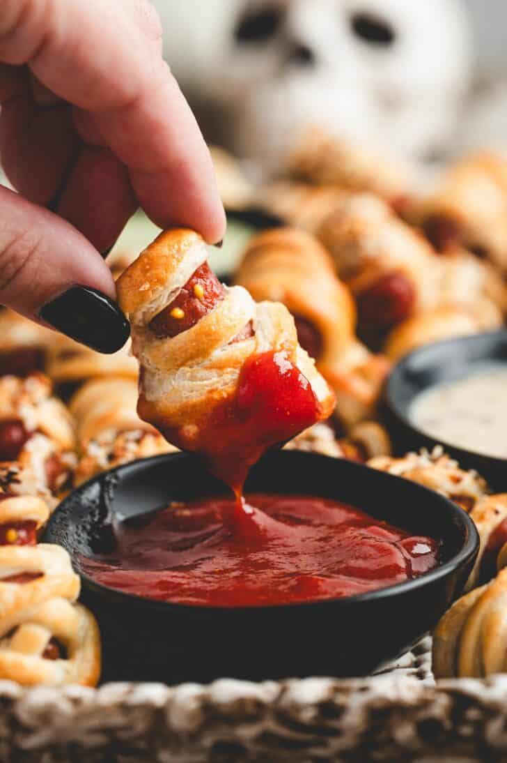 Close-up of a little smokies pig in a blanket being dipped into ketchup, with more mummies blurred in the background