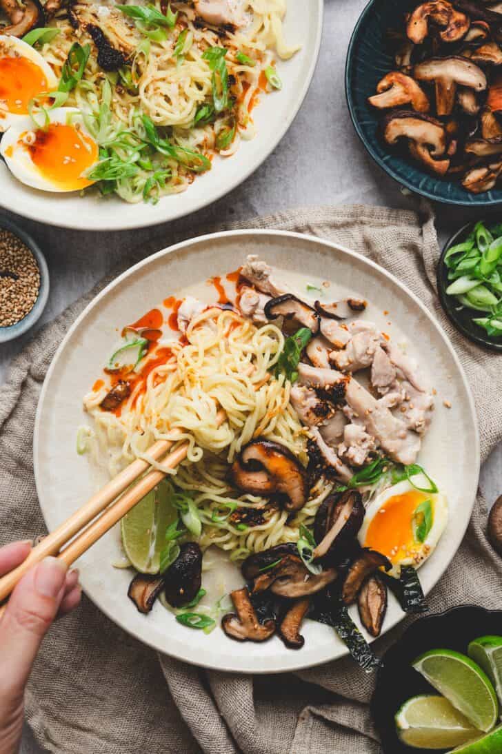 Overhead shot of miso ramen recipe being eaten with chopsticks, surrounded by lime wedges and a bowl of sautéed mushrooms