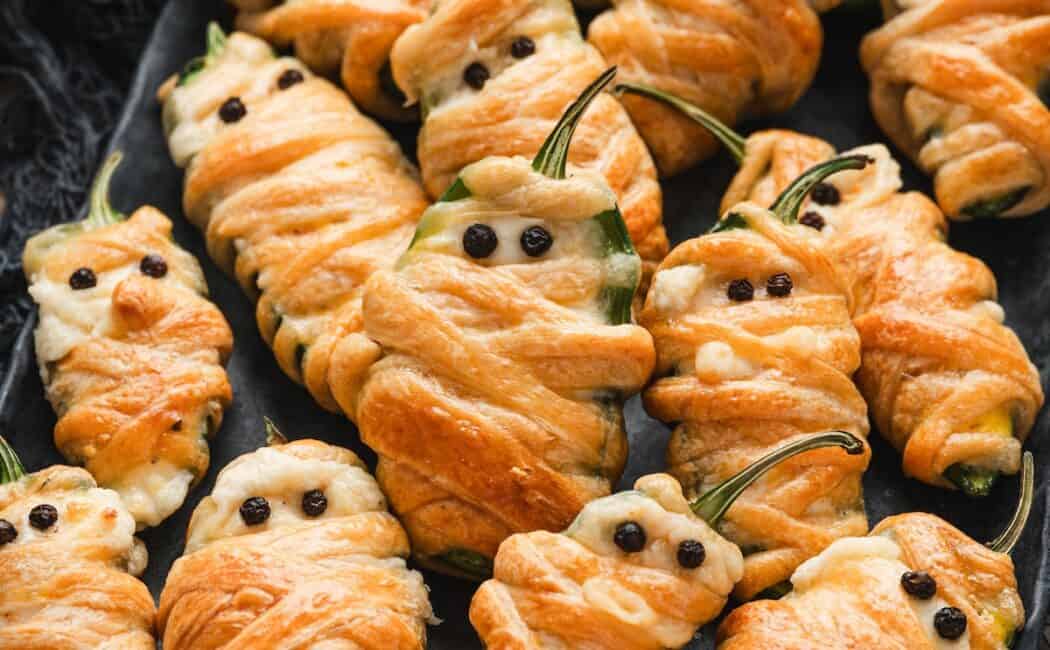Close-up of mummy jalapeno poppers wrapped in golden pastry with peppercorn eyes, piled together on a dark platter.