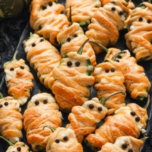 Close-up of mummy jalapeno poppers wrapped in golden pastry with peppercorn eyes, piled together on a dark platter.