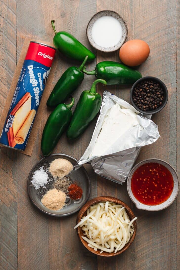 Flat-lay of ingredients for mummy jalapeño poppers, including whole jalapeño peppers, a package of crescent dough sheet, cream cheese, shredded Monterey Jack cheese, an egg, milk, black peppercorns, garlic powder, onion powder, salt, and paprika, with a bowl of sweet chili sauce.