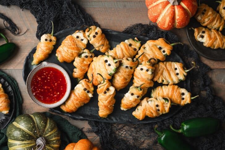 Overhead view of a platter of mummy jalapeno poppers recipe, wrapped in golden pastry with peppercorn eyes, served with a bowl of sweet chili sauce and surrounded by mini pumpkins and jalapeños.