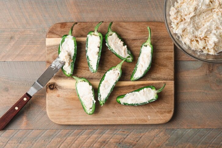 Overhead view of jalapeño pepper halves filled with a cream cheese and Monterey Jack mixture on a wooden cutting board, with a knife and bowl of filling alongside.