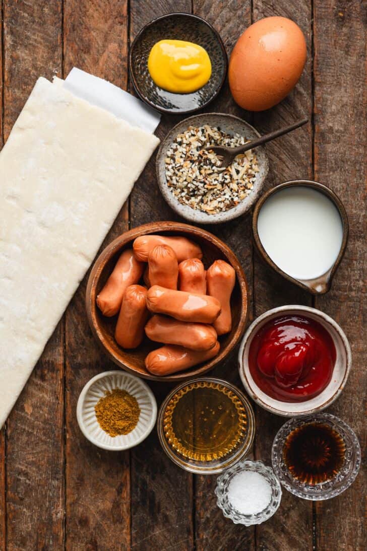 Ingredients for mummy pigs in a blanket laid out, including puff pastry, little smokies sausages, mustard, egg, milk, ketchup, curry powder, and everything bagel seasoning