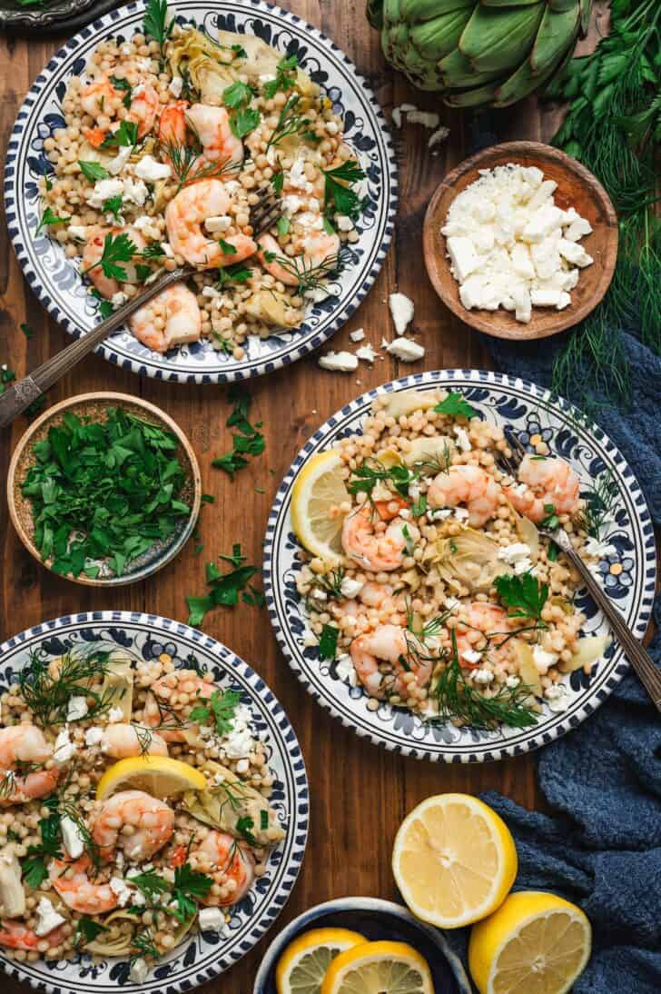 Overhead view of three plates of a pearl couscous recipe topped with shrimp, artichokes, crumbled feta, dill, and parsley, served with lemon wedges, a bowl of chopped parsley, and a bowl of feta alongside.