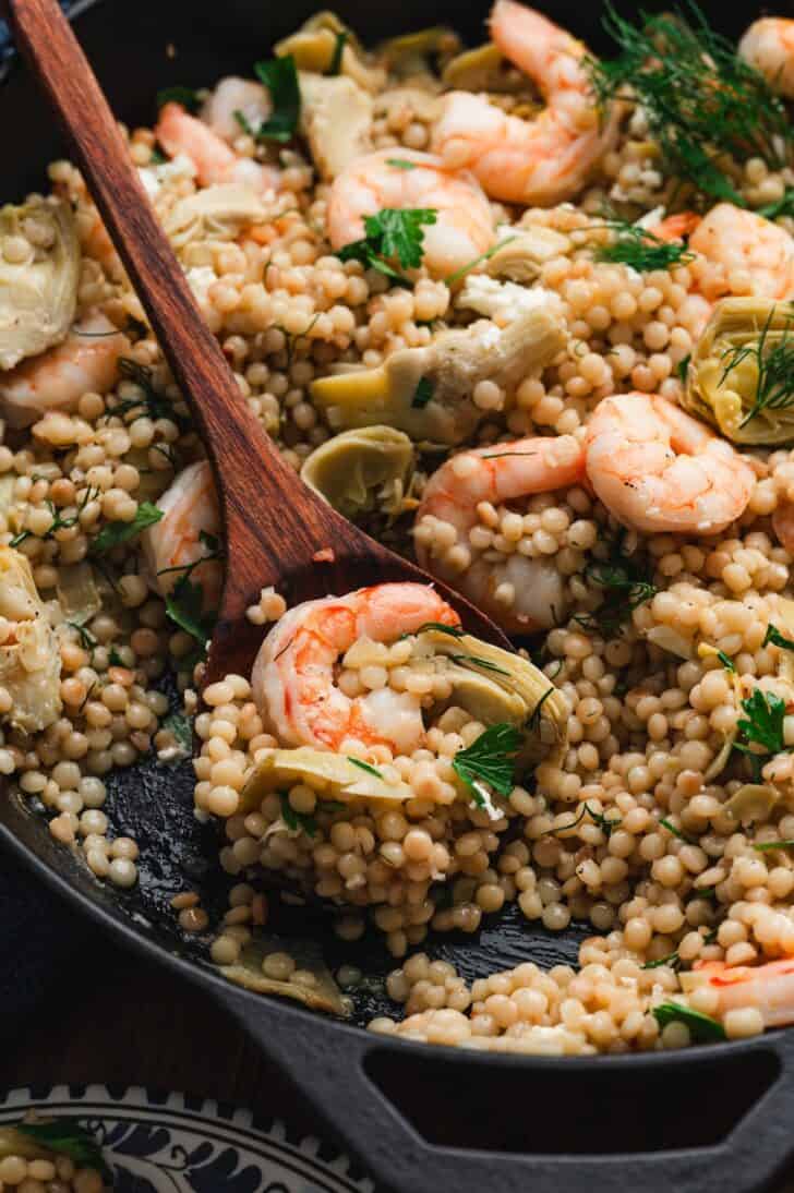 Close-up of pearl couscous with shrimp in a cast iron skillet, tossed with artichokes, crumbled feta, fresh dill, and parsley, with a wooden spoon resting in the dish.