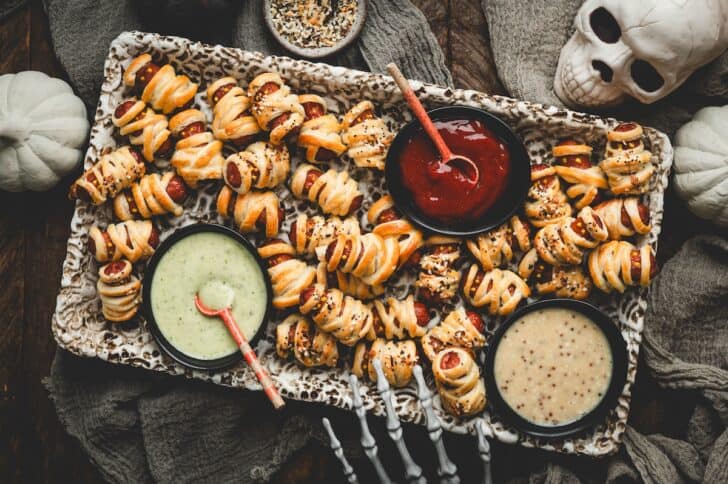 Wide overhead shot of a platter of pigs in a blanket puff pastry mummies with dipping sauces, a skull, and white pumpkins