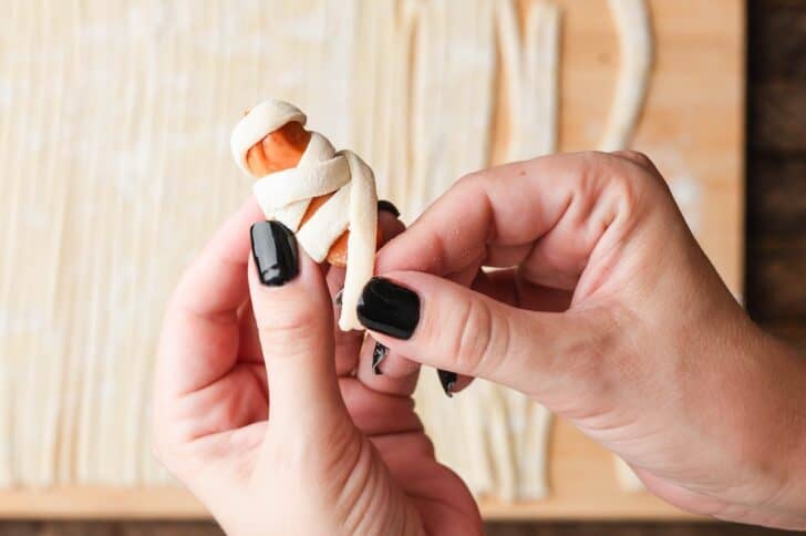 Hands wrapping a strip of dough around a cocktail sausage to shape pigs in a blanket puff pastry mummies