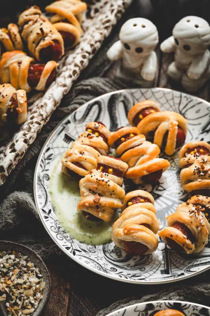 Plate of puff pastry pigs in a blanket served with a pesto ranch dip, with mummy-shaped salt and pepper shakers nearby