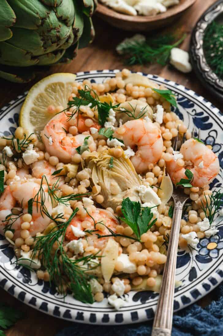 Close-up of a plated shrimp artichoke recipe with pearl couscous, garnished with crumbled feta, fresh dill, parsley, and a lemon wedge.