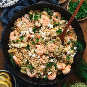 Overhead view of a shrimp couscous skillet with artichokes, crumbled feta, fresh dill, and parsley, served with lemon slices and a bowl of chopped parsley alongside.