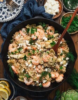 Overhead vertical view of a shrimp couscous recipe in a cast iron skillet, topped with crumbled feta, fresh dill, and parsley, served with lemon slices, a bowl of feta, and a bowl of dill alongside.