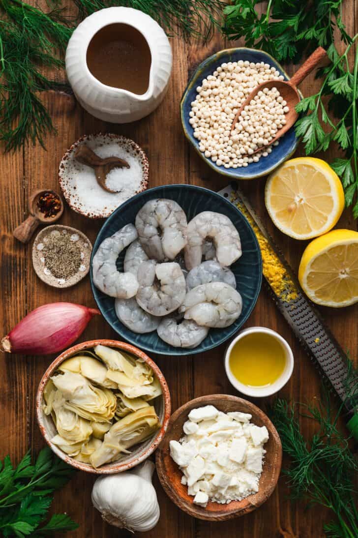 Flat-lay of ingredients for shrimp couscous, including peeled and deveined shrimp, pearl couscous, canned artichoke quarters, crumbled feta, a shallot, garlic, chicken stock, olive oil, lemon halves, salt, pepper, red pepper flakes, and fresh dill and parsley.