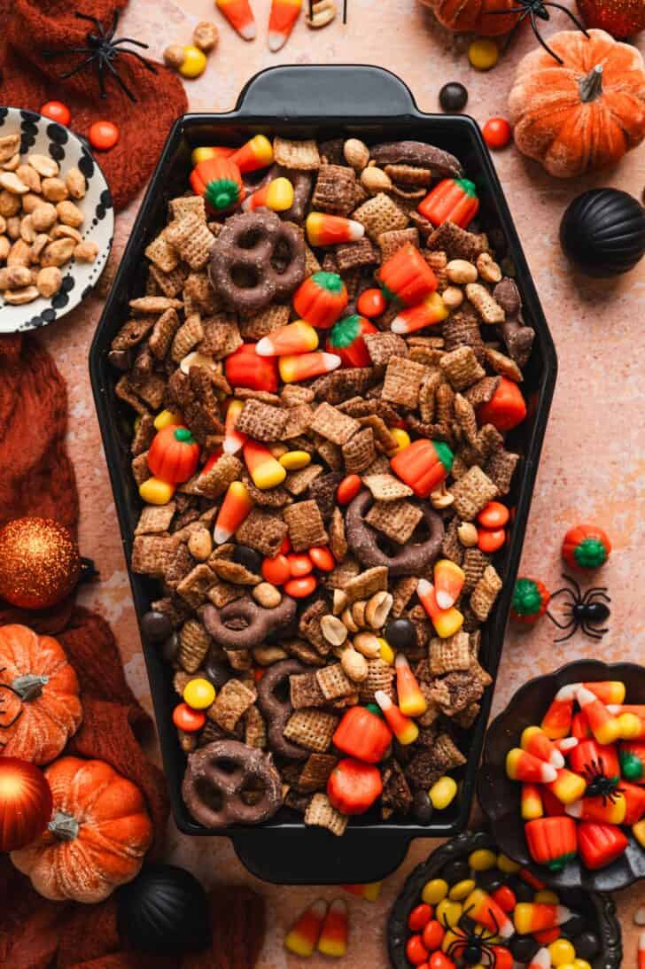 Coffin-shaped dish filled with sweet chex mix, surrounded by Halloween decorations like pumpkins and spiders