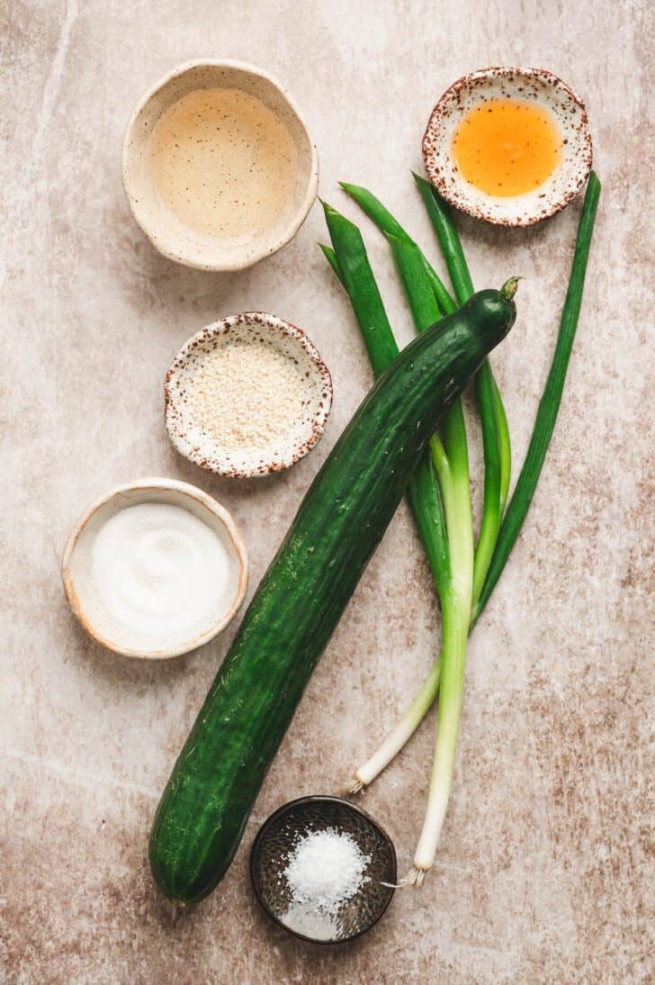 Ingredients for the salad laid out, including a whole English cucumber, green onions, rice vinegar, sugar, sesame seeds, sesame oil, and kosher salt